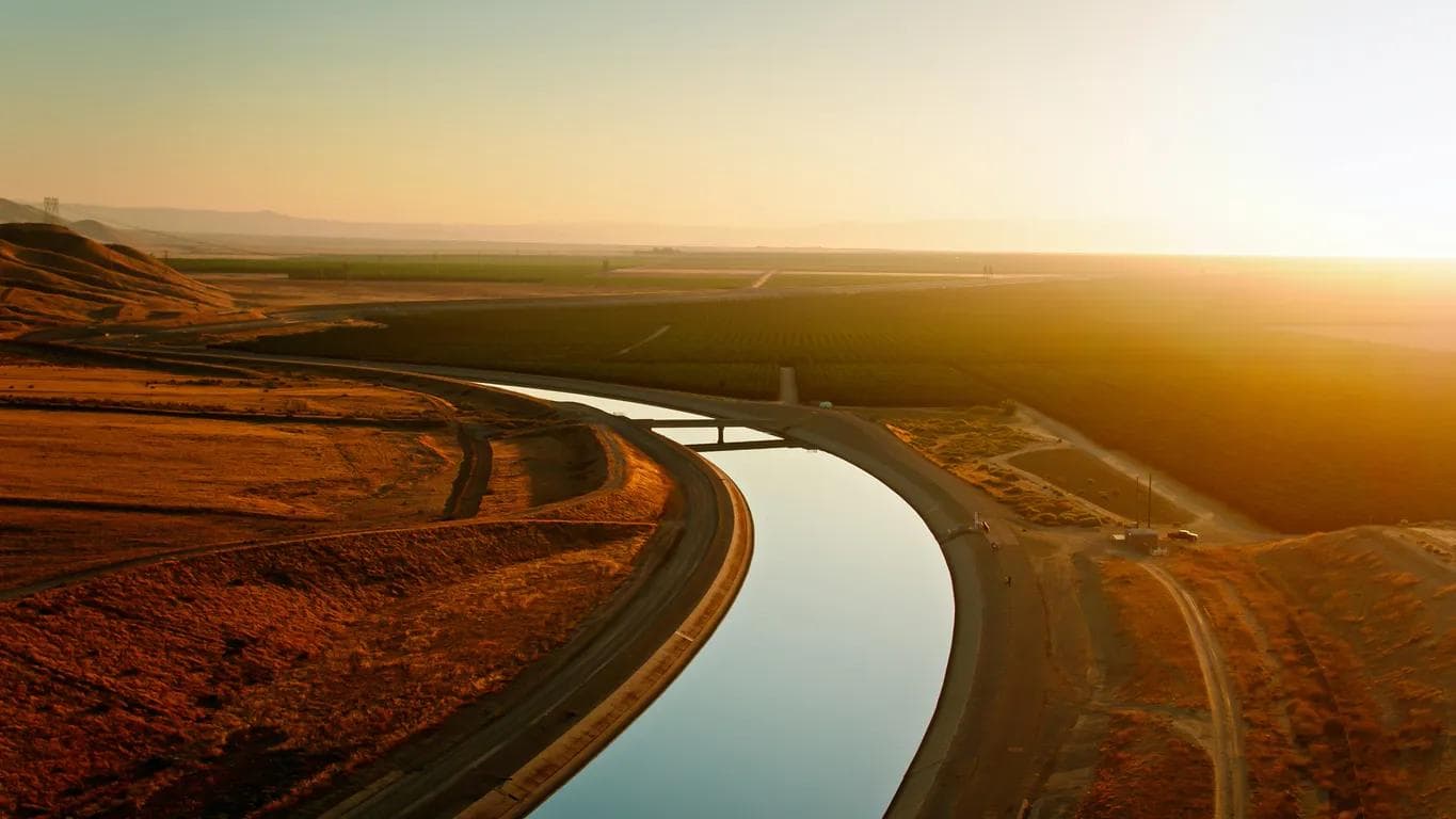 Aerial view of the California Aqueduct curving between a barren hillside and lush agricultural fields in Kern County, California