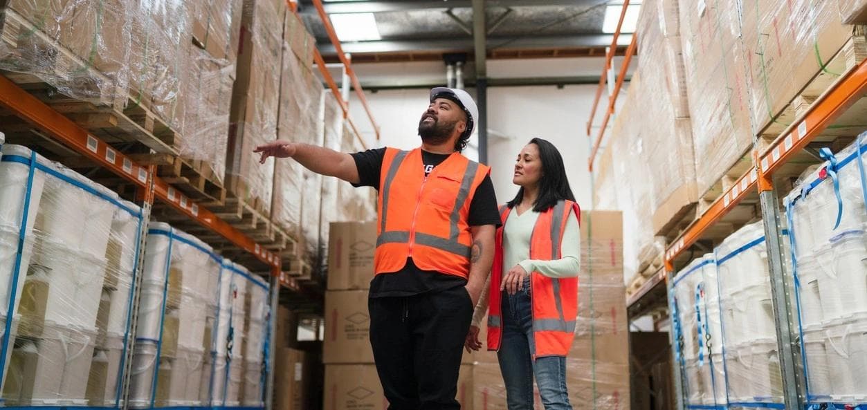 Two logistics workers in reflective safety gear review shipment details while walking through a busy warehouse facility.